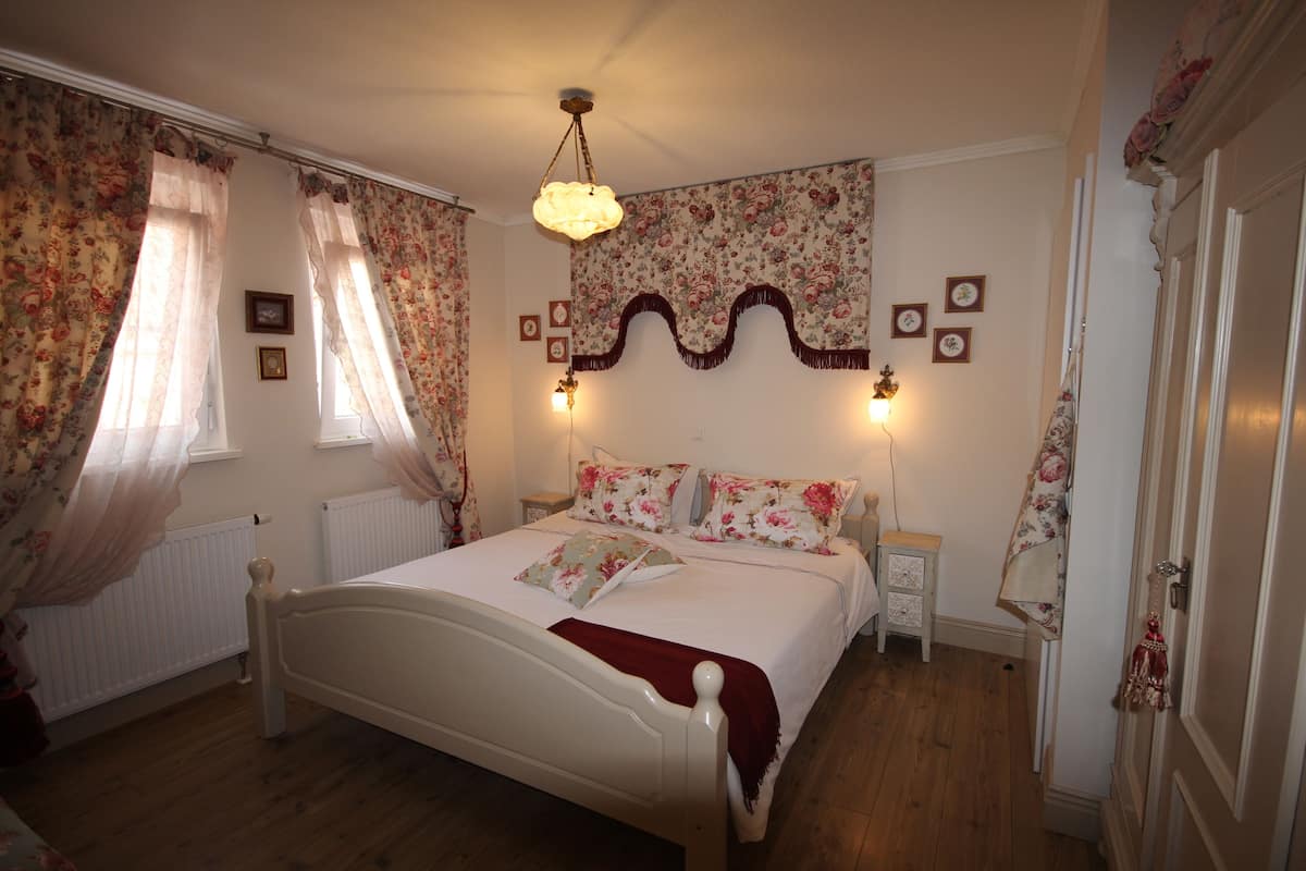 A bedroom with a white bed, floral curtains, a wooden floor, and a decorative wall hanging.