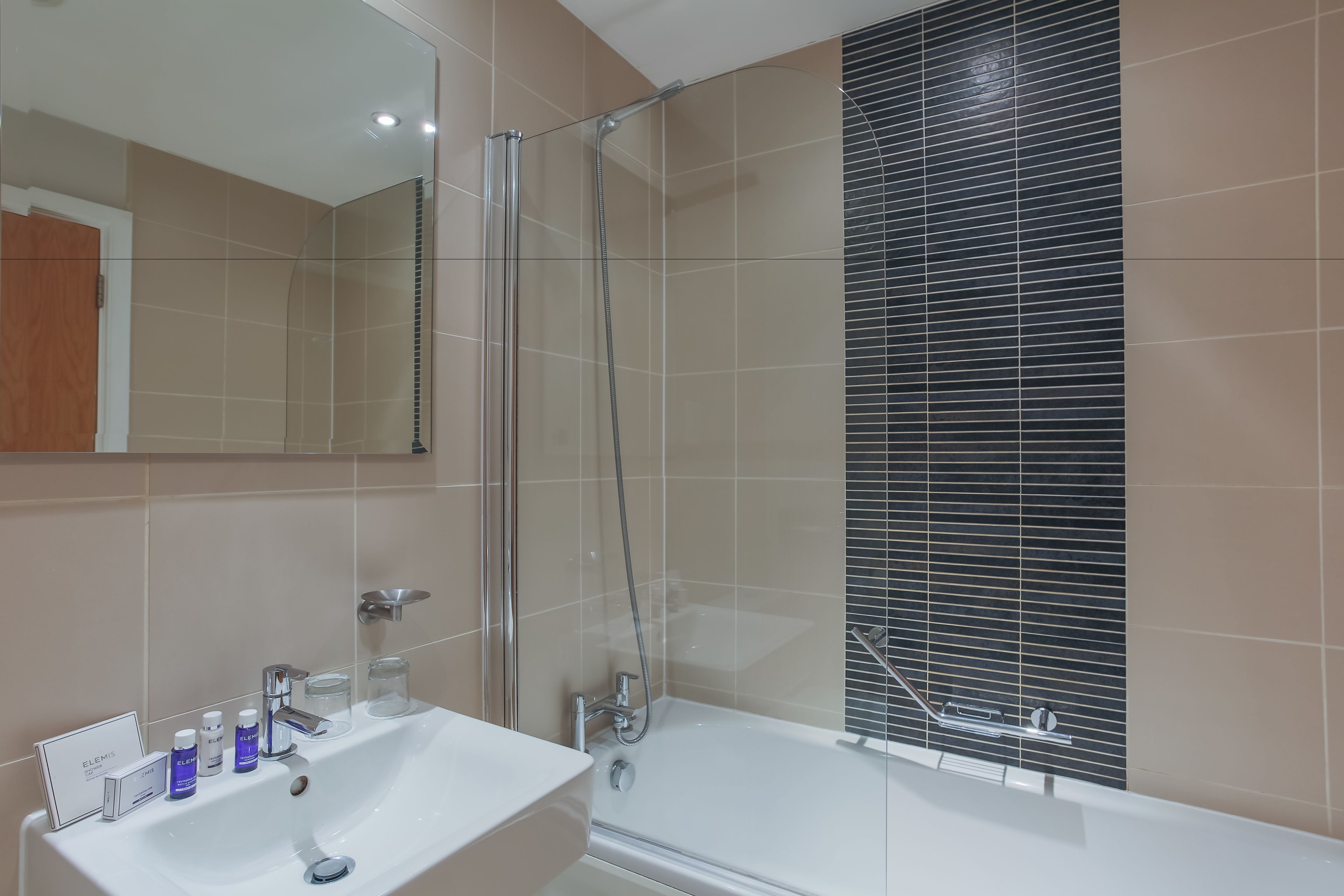 Un baño moderno con ducha de vidrio, un lavabo blanco con artículos de aseo y un espejo grande.