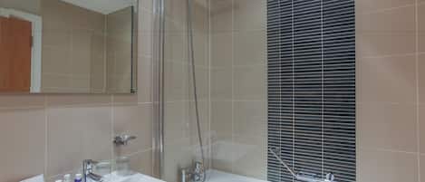 Un baño moderno con ducha de vidrio, un lavabo blanco con artículos de aseo y un espejo grande.