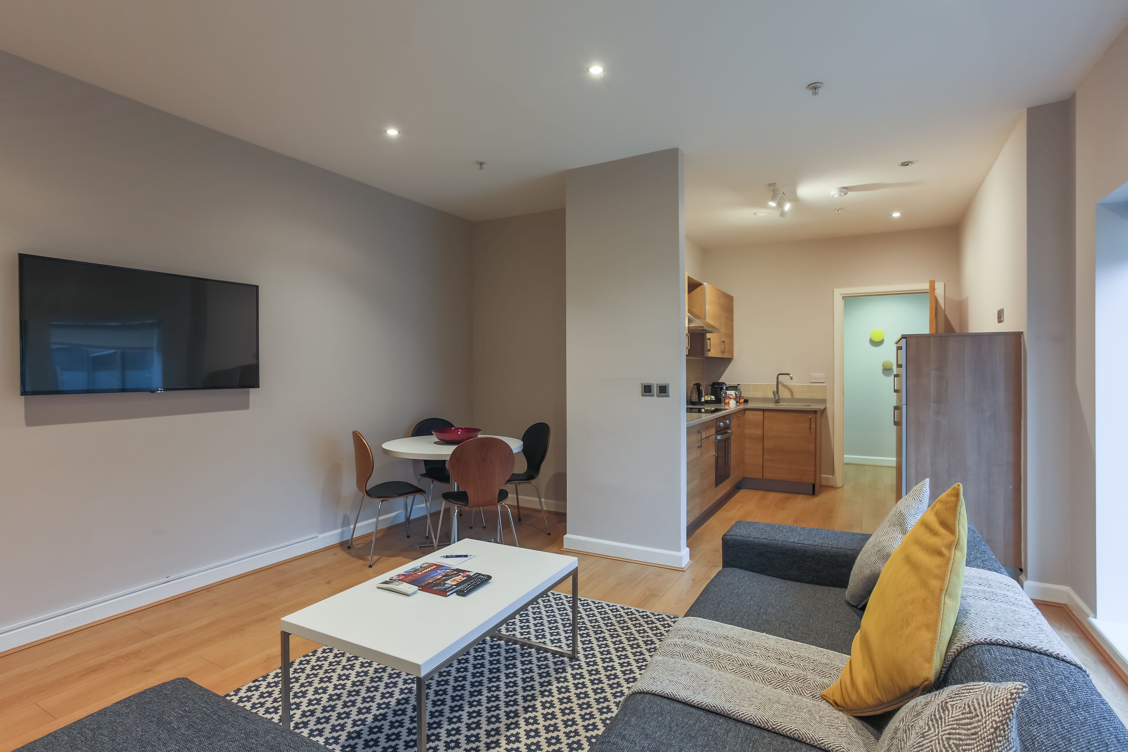 A modern living room with a grey sofa, a white coffee table, and a patterned rug. There is a dining area with a table and chairs, and a kitchen in the background.