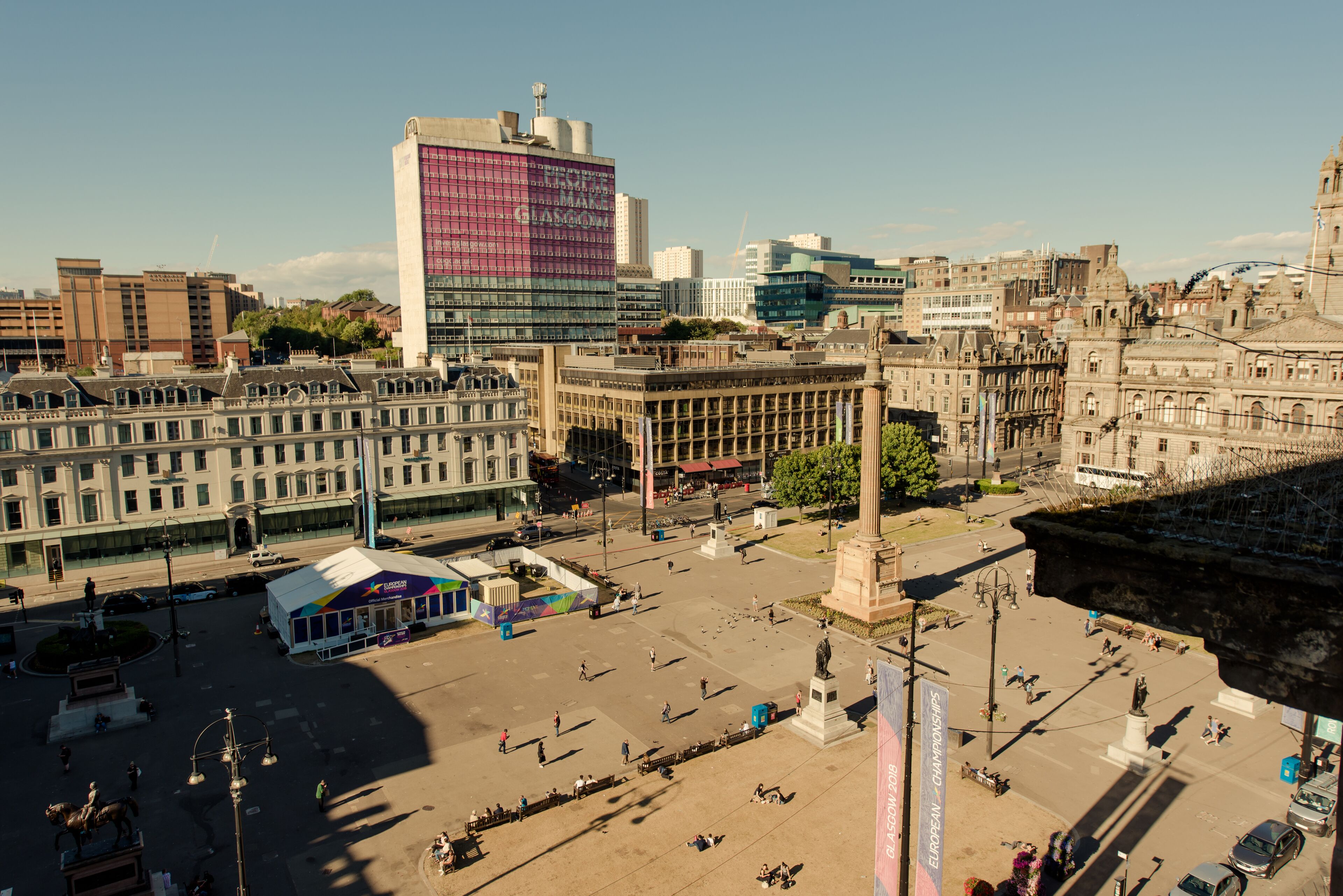 Uma praça urbana com um monumento, edifícios e um amplo edifício moderno com fachada rosa e branca.