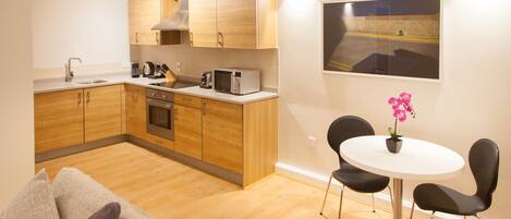 Een moderne keuken met houten kastjes, een inbouwoven en een magnetron. Er is een eethoek met een witte tafel en zwarte stoelen.