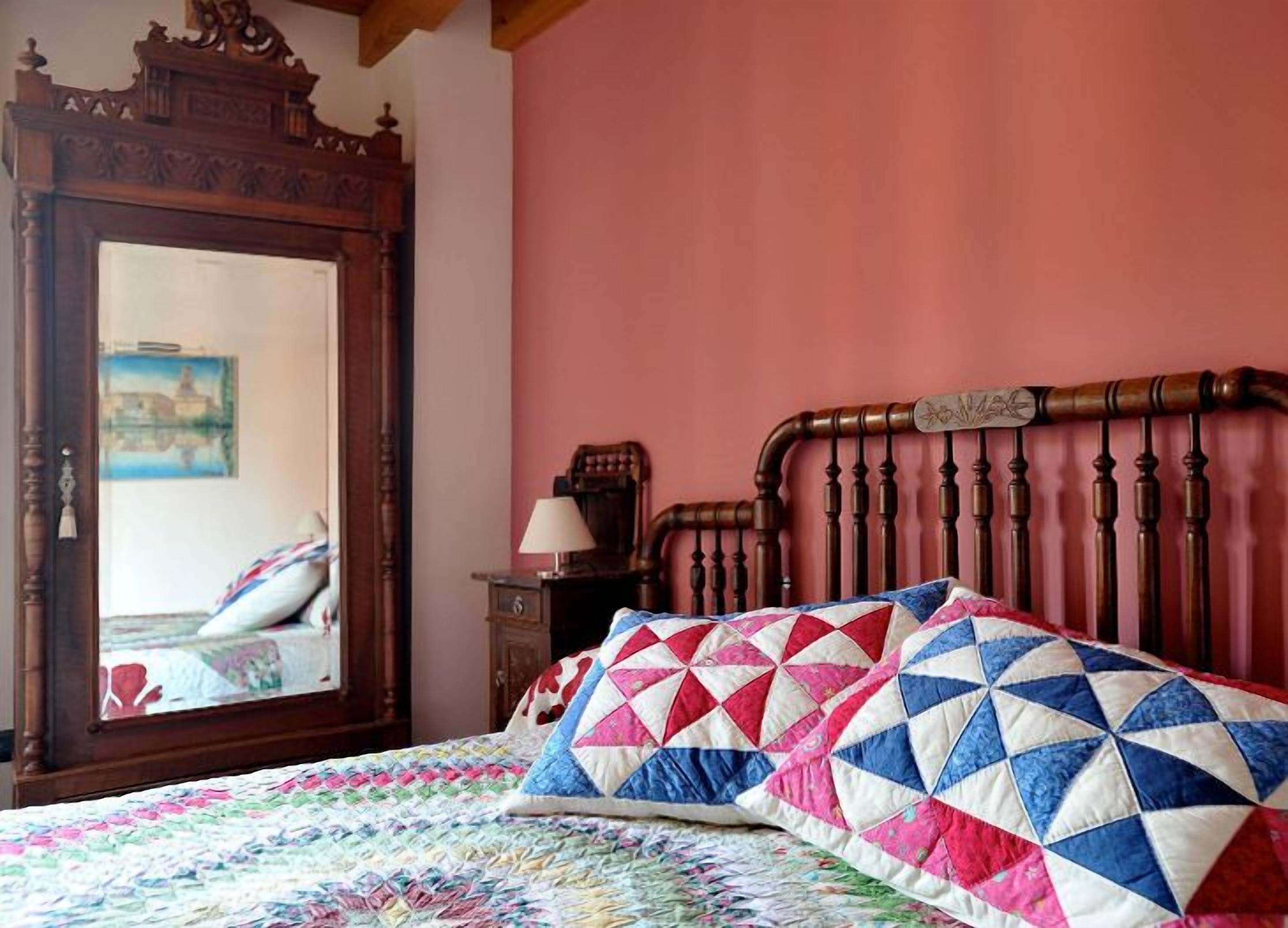 Un dormitorio con un cabecero de madera, colcha colorida, espejo y mesita de noche con lámpara.