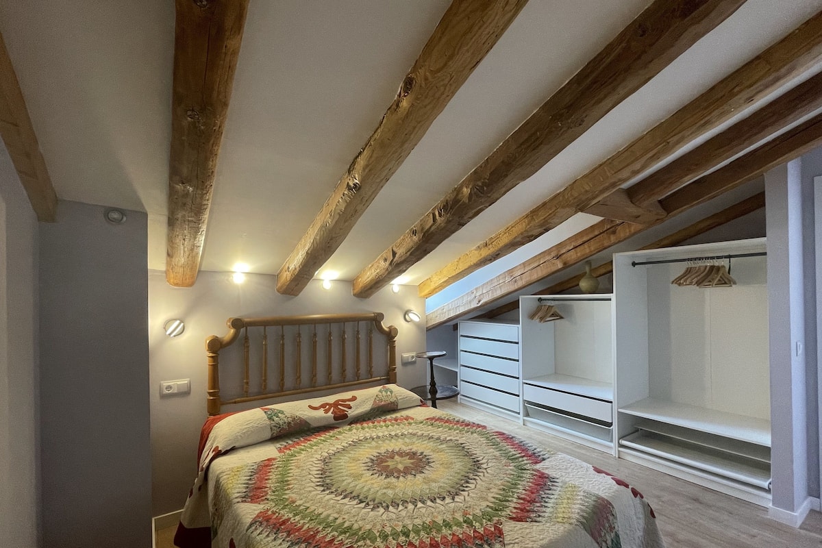 Une chambre avec un plafond en bois, un lit recouvert d’une courtepointe à motifs, une commode blanche et un dressing.