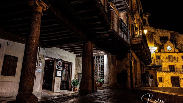 Vista nocturna de un edificio histórico con galería techada, columnas de piedra y torre del reloj.