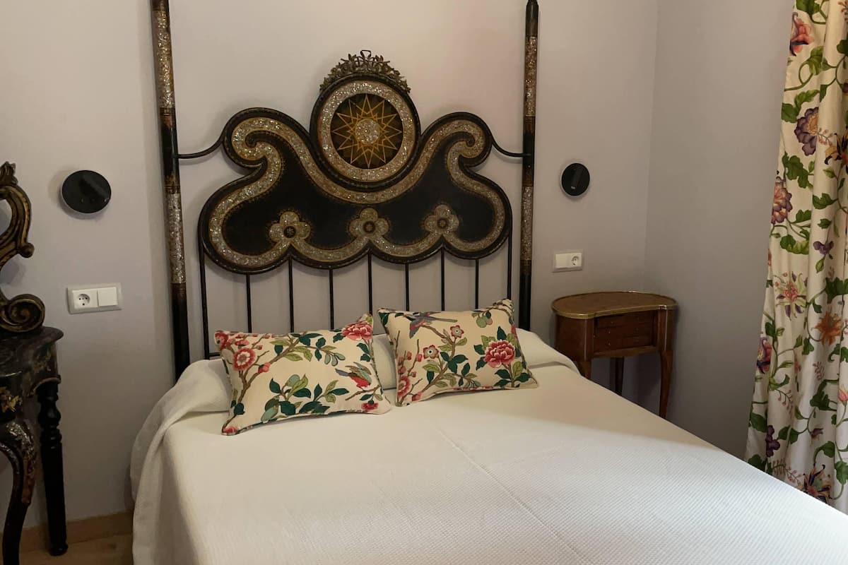 Un lit agrémenté de coussins à motifs floraux, d’une tête de lit décorative, d’une table de chevet en bois et d’un rideau à fleurs.