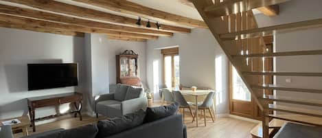 A modern living room with a wooden ceiling, a flat-screen TV, a grey sofa, and a dining area with a table and chairs.