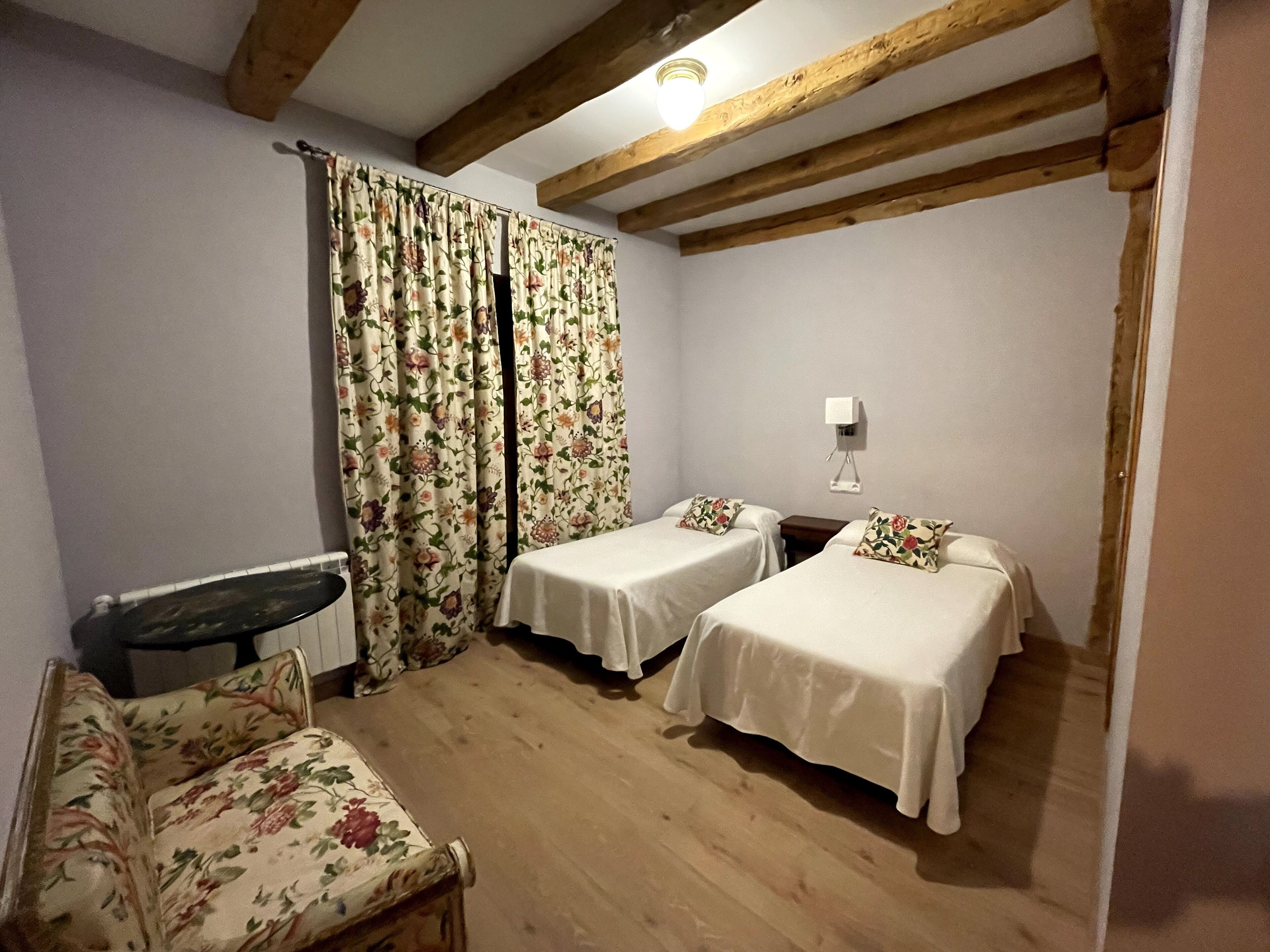 Una habitación con dos camas individuales, techo de madera, una mesita pequeña y una silla con tapicería floral.