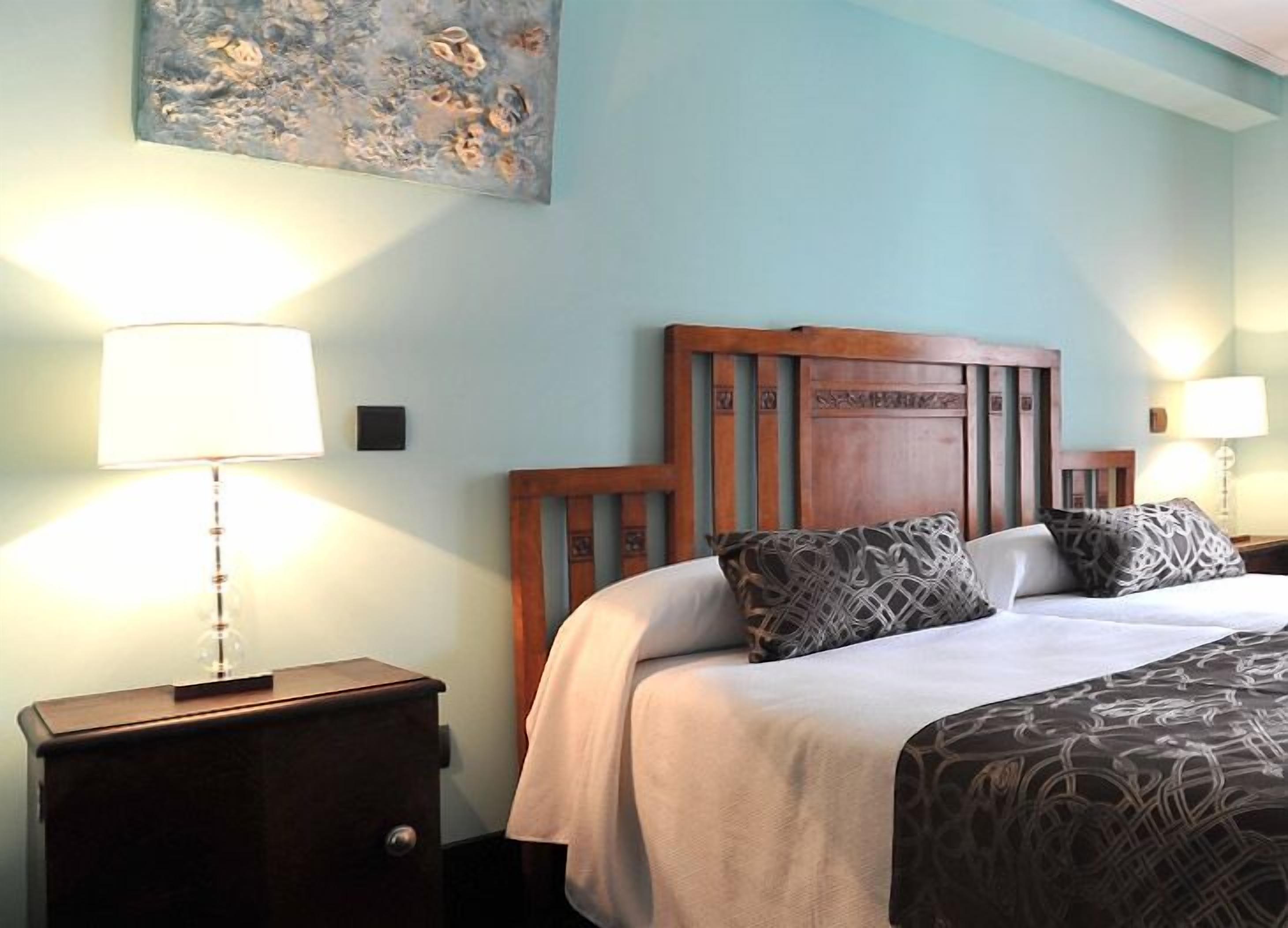 Habitación de hotel con cabecera de madera, dos mesitas de noche con lámparas, cama con ropa de cama estampada y un cuadro en la pared.