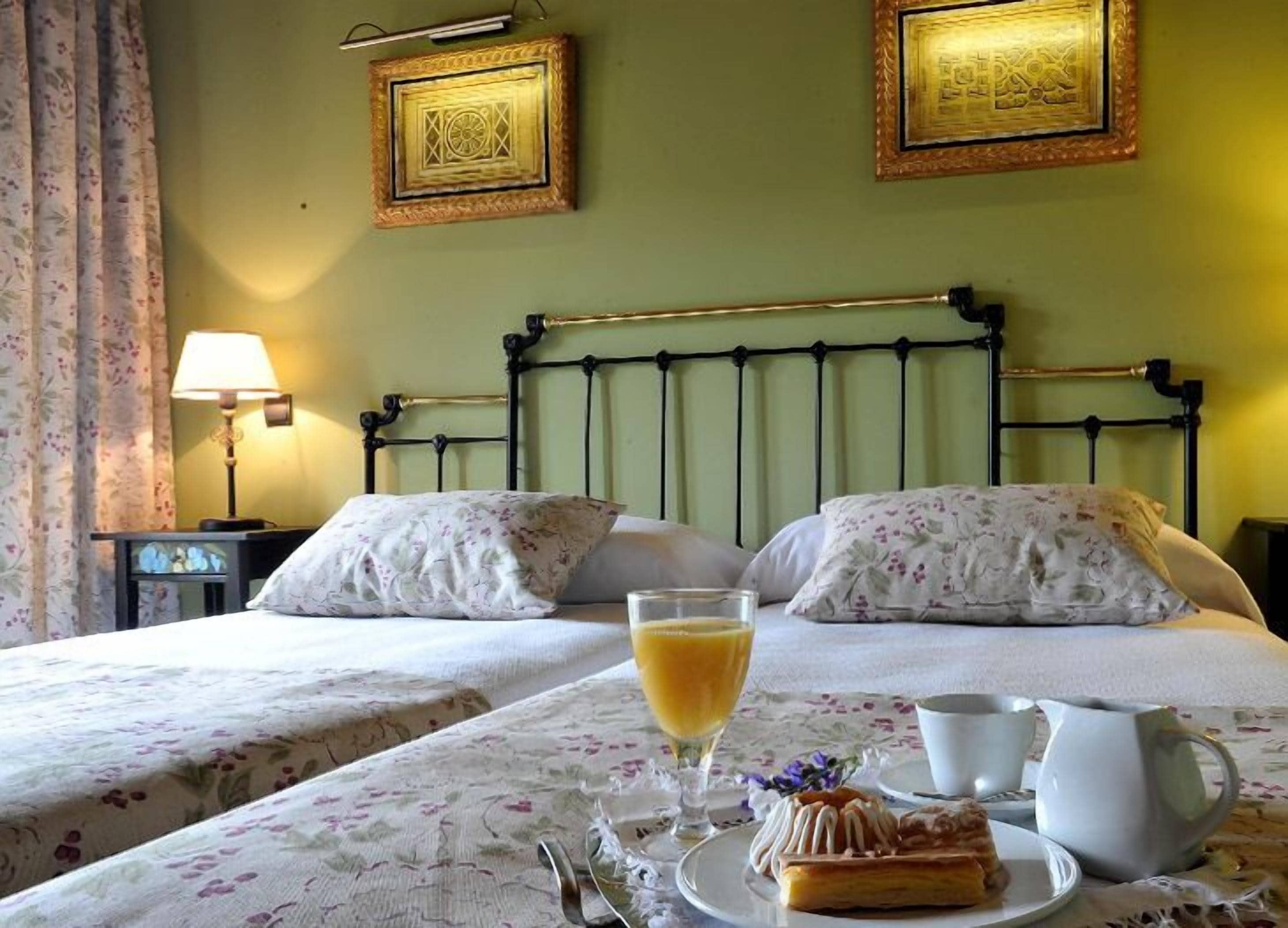 A hotel room with two beds, floral bedding, a bedside table with a lamp, and a breakfast setup with a glass of orange juice, a pastry, and a pitcher.
