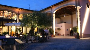 An outdoor patio area with tables and chairs, a large tree, and a building with arches and a covered area. - Agriturismo Argaland (Parma)