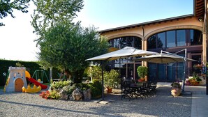 A playground with a slide and swings, a covered seating area with tables and chairs, and a building with large windows and a curved facade. - Agriturismo Argaland (Parma)