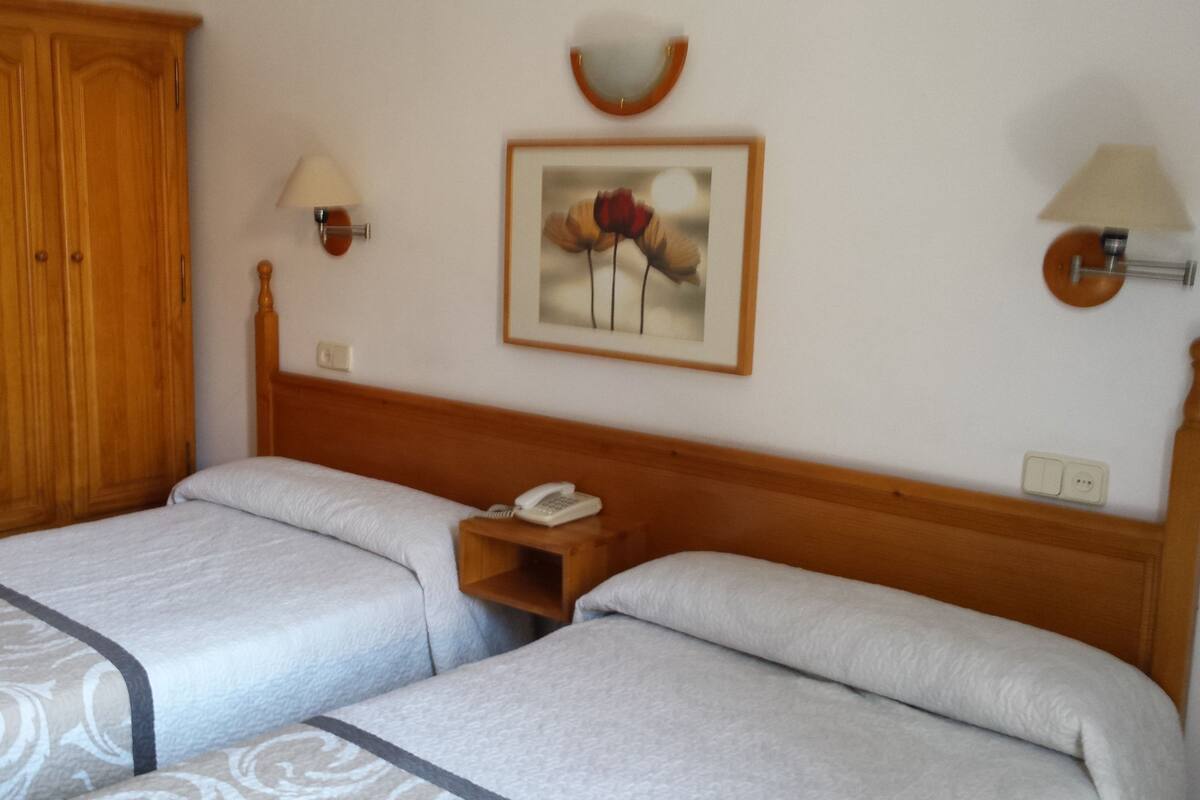 Habitación doble con dos camas, un armario de madera, una mesita de noche con lámpara y un cuadro en la pared.