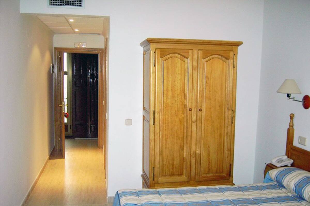 Un dormitorio con un armario de madera, una cama con colcha a rayas y una mesita de noche con lámpara.