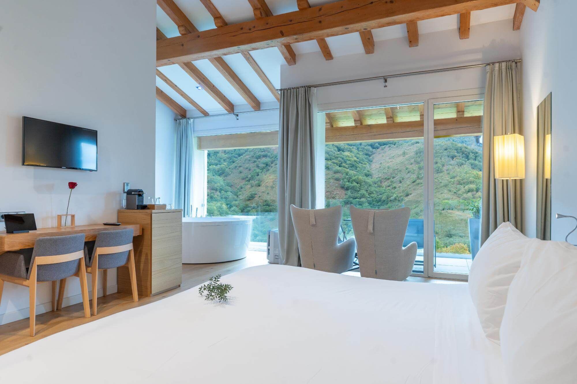 A bedroom with a bed, a desk with chairs, a TV, and a view of a forested area.