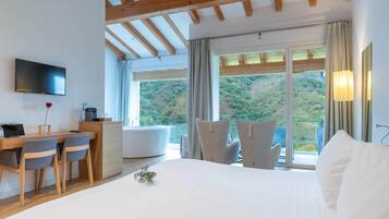A bedroom with a bed, a desk with chairs, a TV, and a view of a forested area.
