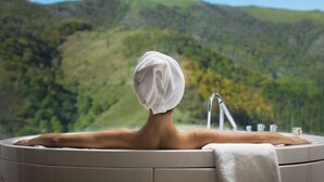 Eine Person sitzt in einem Whirlpool, mit einem Handtuch um den Kopf gewickelt, vor einer Berglandschaft.