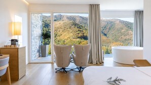 Ein Hotelzimmer mit Bett, zwei Sessel, Schreibtisch, Lampe, einem großen Fenster mit Blick auf eine gebirgige Landschaft und einer Badewanne.