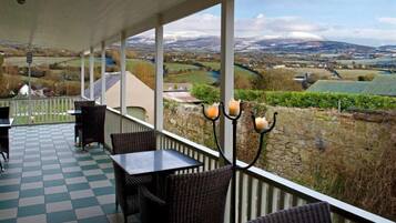 A covered patio with checkered flooring, wicker furniture, and a view of a rural landscape with fields and mountains.