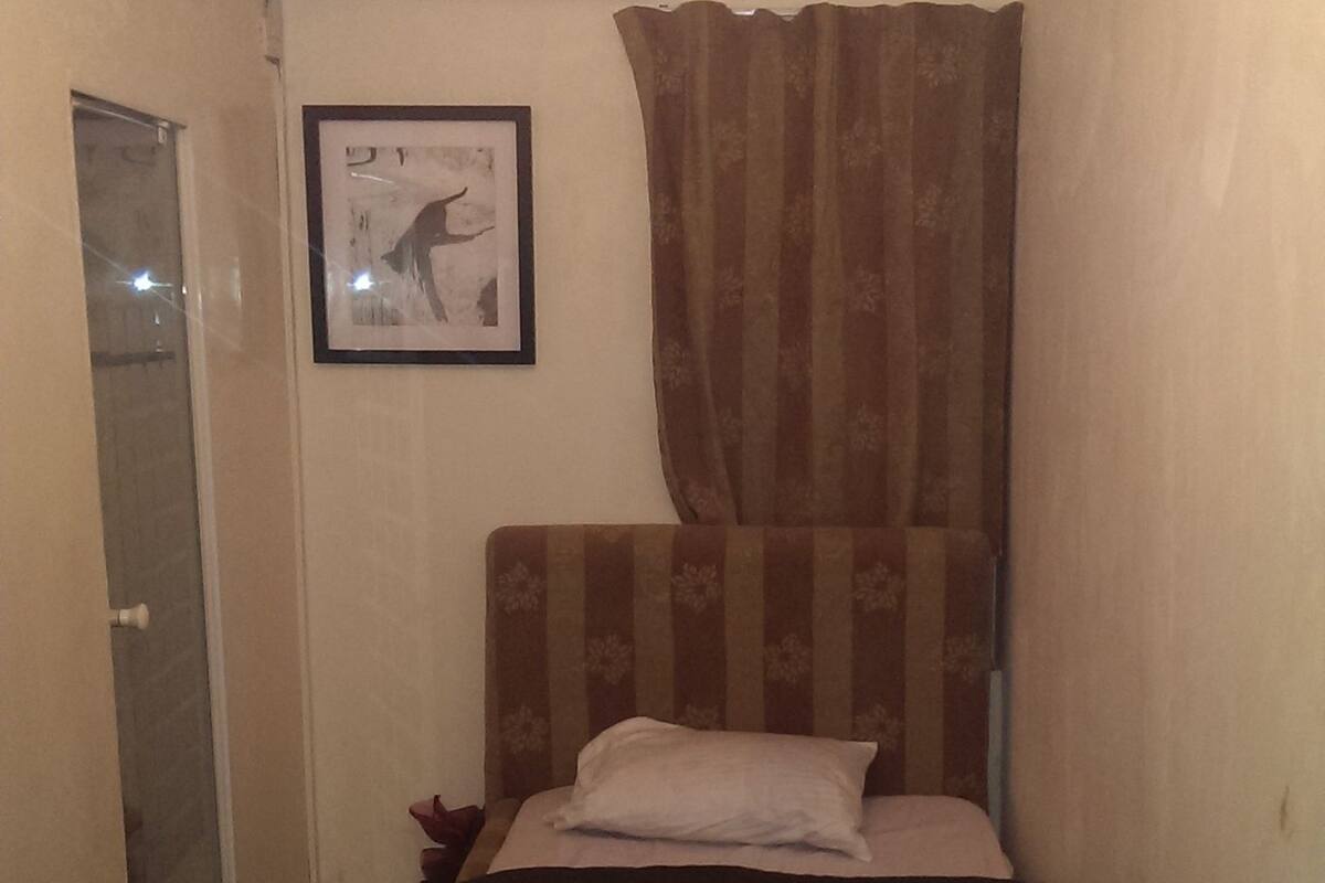 Una habitación pequeña y estrecha con una cama, una ventana con cortinas, un equipo de aire acondicionado y un cuadro en la pared.