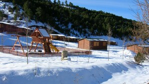 A snow-covered outdoor area with a wooden swing set, a fenced play area, and several wooden cabins in a mountainous setting. - Cabañas de Javalambre (Camarena de la Sierra)