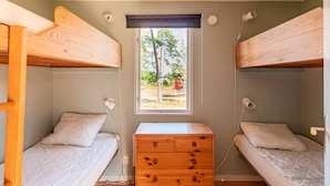 Une petite pièce compacte avec deux lits superposés, une commode en bois avec des tiroirs, une fenêtre donnant sur des arbres et un objet rouge à l’extérieur, ainsi qu’une prise électrique sur le mur.