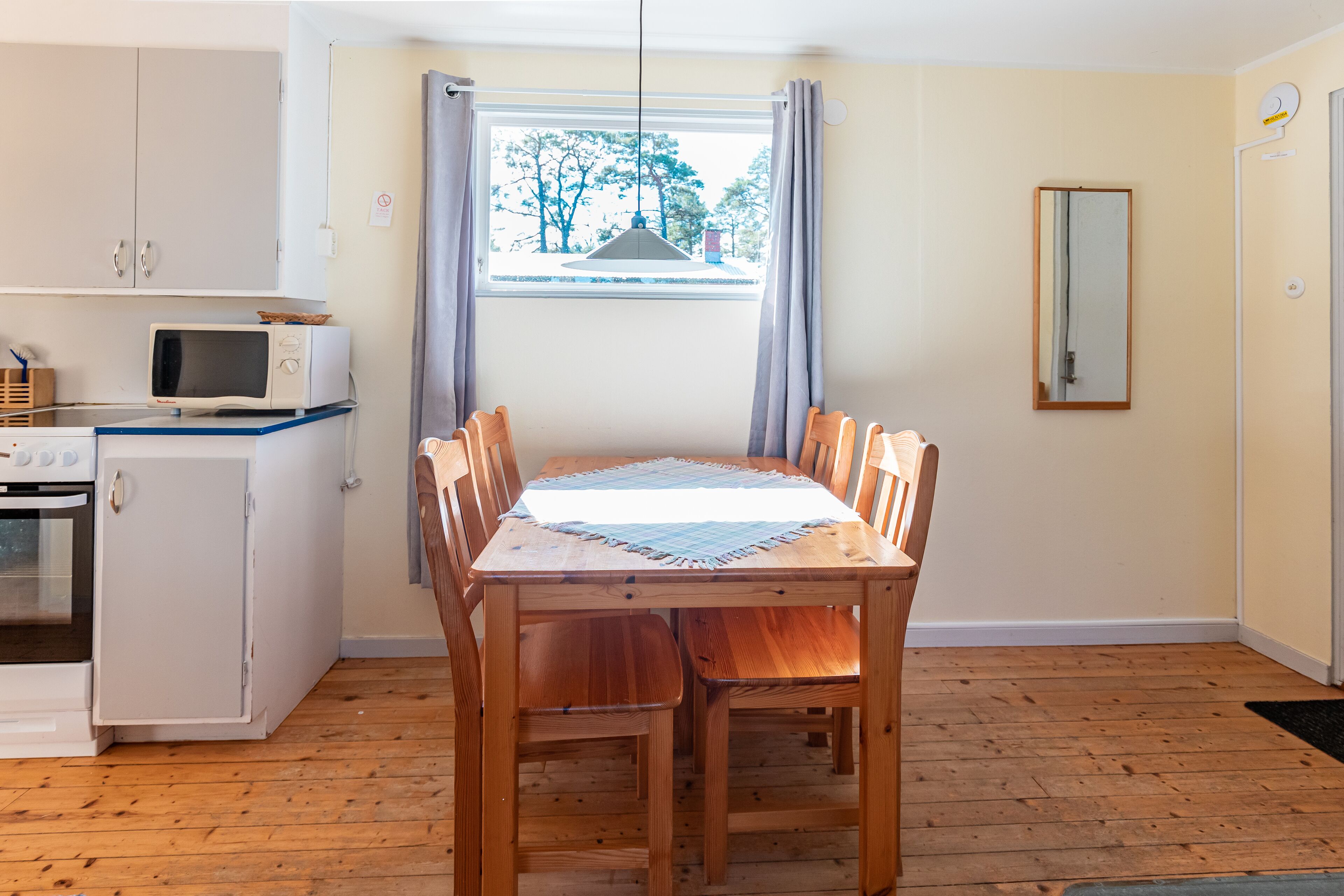 A compact kitchen with a microwave, oven, and a small refrigerator. There is a dining table with chairs and a window with curtains.