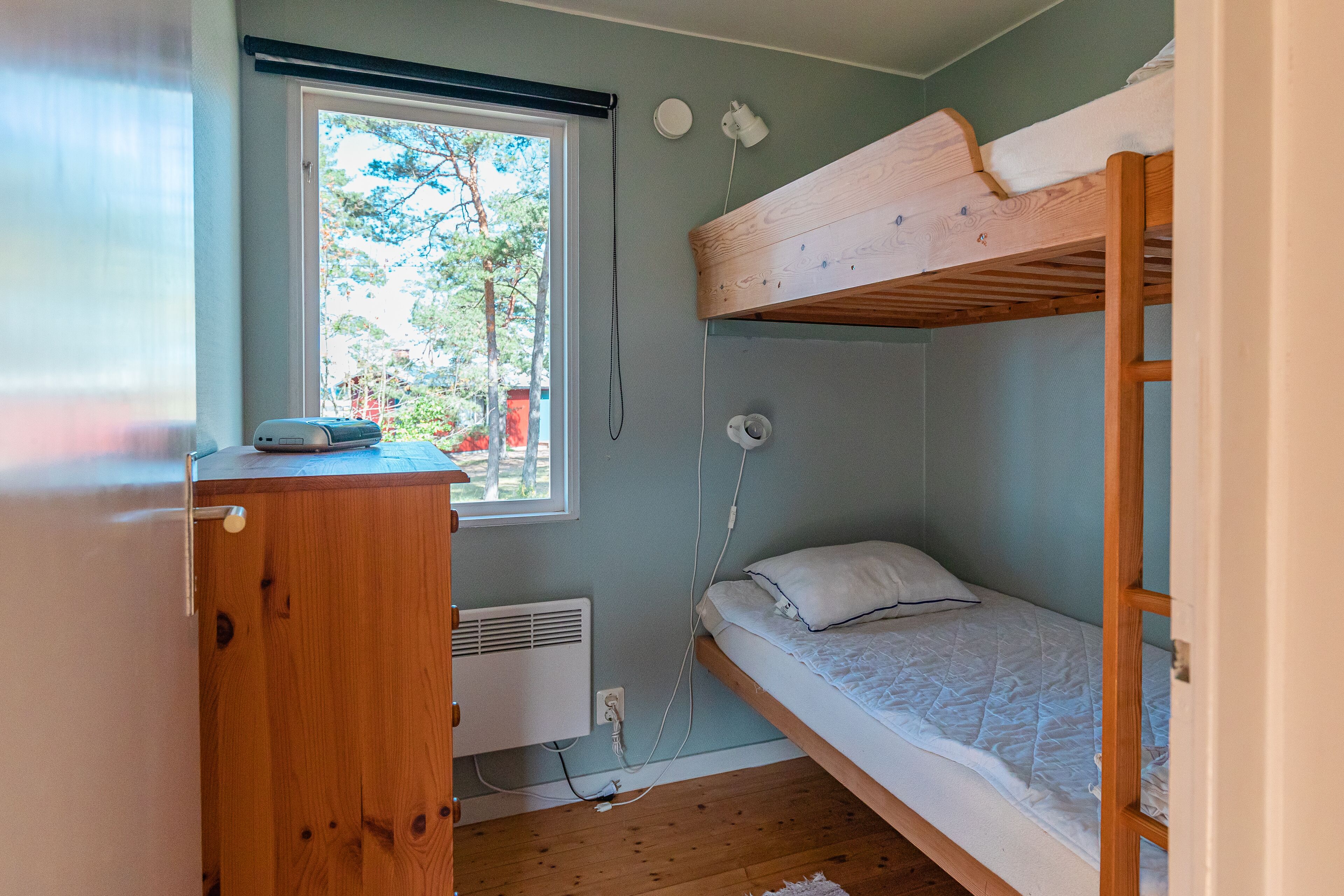 A small, compact room with a bunk bed, a wooden desk, a window with a view of trees, and a wall-mounted heater.