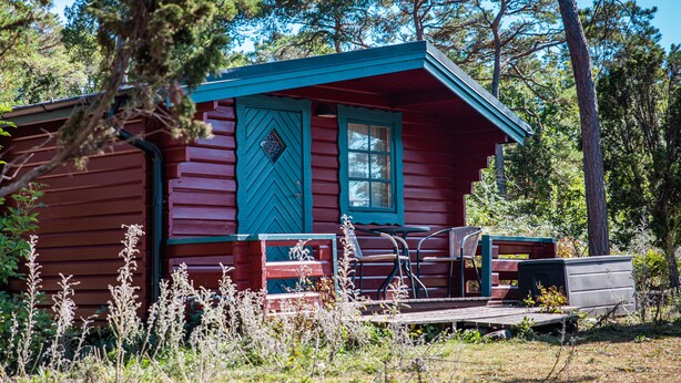 Une cabane à l’extérieur rouge et aux bordures bleues, une véranda avec un banc et un petit abri de jardin.