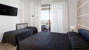 A hotel room with two beds, a TV, a window with curtains, and a view of the city. - Residenza Levante (Riccione)