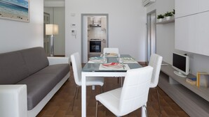 A modern living room with a glass dining table, white chairs, a grey sofa, a flat-screen TV, and a bookshelf with books. - Residenza Levante (Riccione)