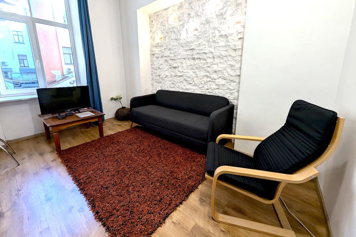 A living room with a black sofa, a wooden coffee table, a TV, a red rug, and a stone wall.