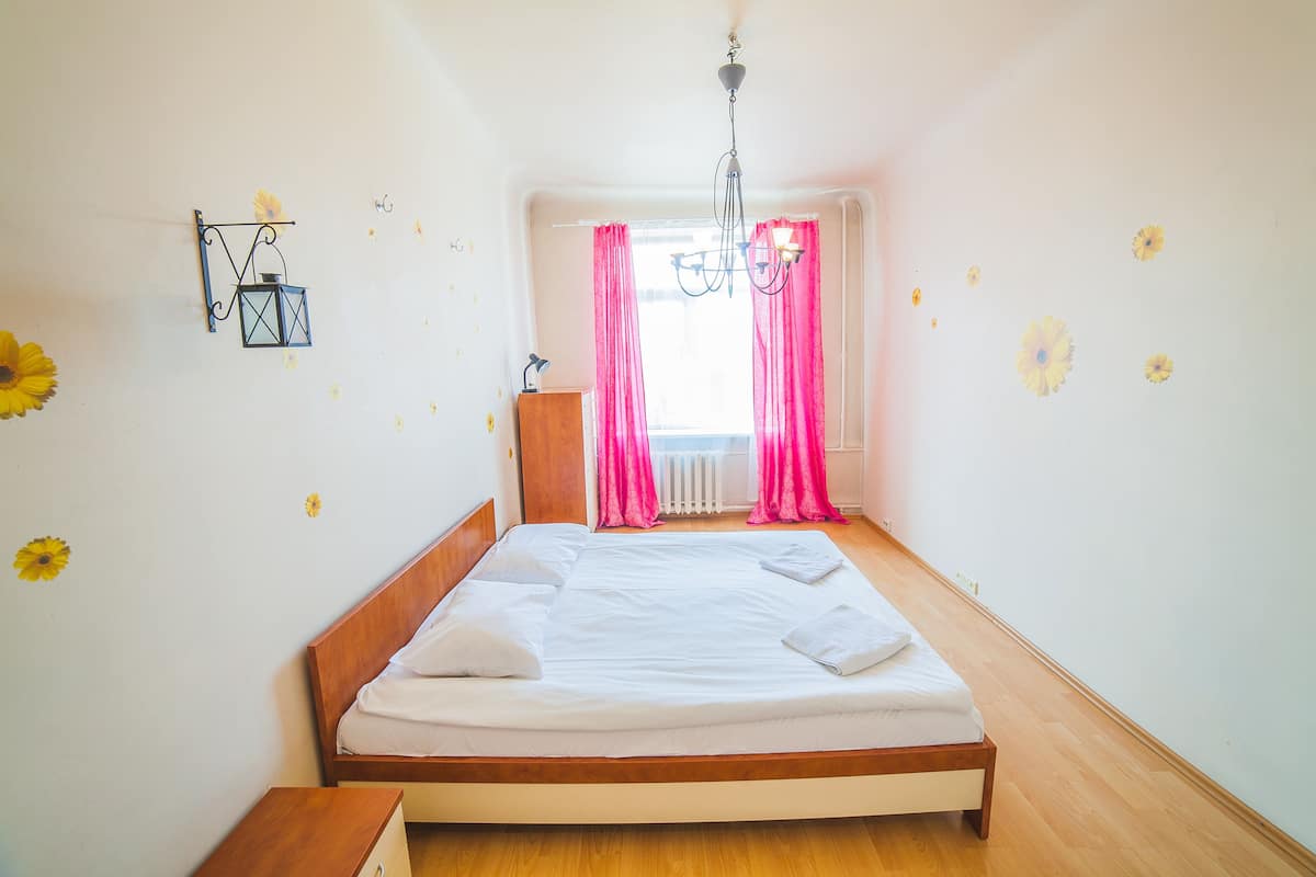 A neatly made bed in a room with a wooden floor, a hanging light fixture, and pink curtains.