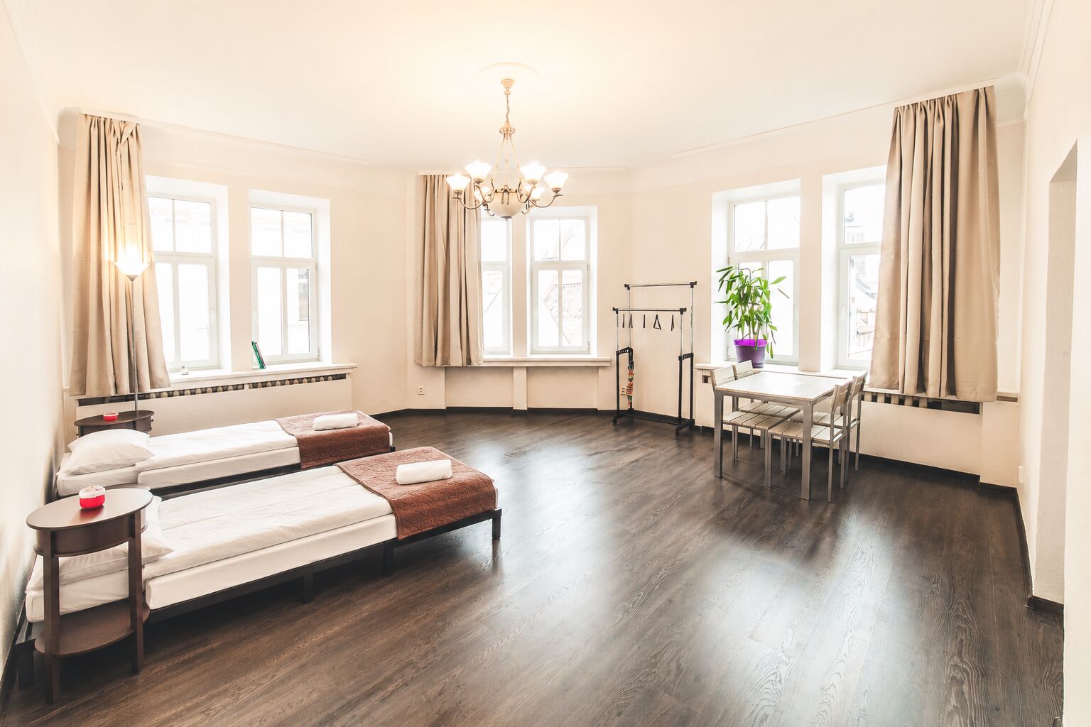 Hotellihuone, jossa on kaksi sänkyä, ruokapöytä, tuoleja, kattokruunu ja suuret ikkunat, joissa on verhot.