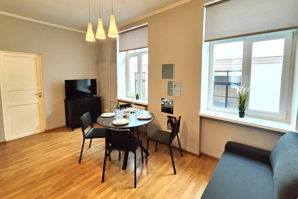A dining area with a round table, black chairs, and hanging pendant lights. There is a black cabinet with a flat-screen TV, a window with a view, and a potted plant on the windowsill.