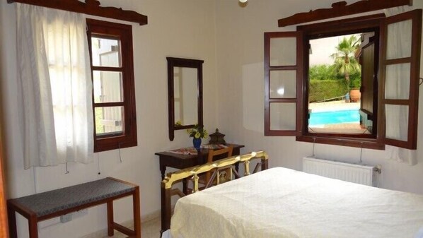 A bedroom with a bed, a bedside table, a chair, a desk with a chair, a window with a view of a pool, and a radiator.