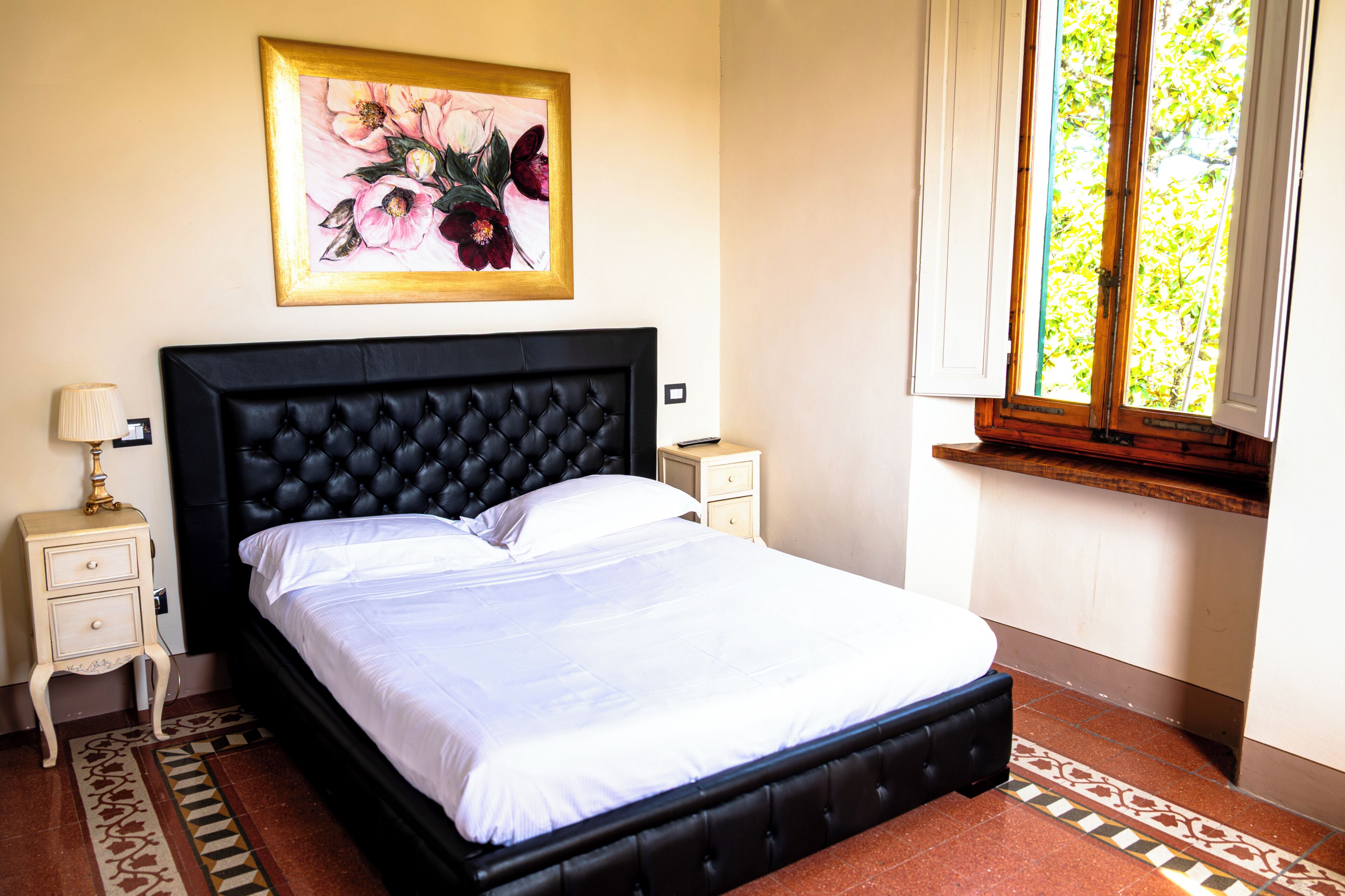 A neatly made bed with a black headboard, a bedside table with a lamp, a framed floral artwork on the wall, and a window with a view of greenery.