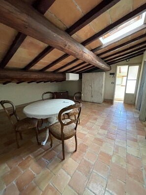 Habitación en ático con una mesa redonda y sillas, vigas de madera a la vista y vistas a través de una puerta abierta.