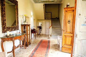 Una imponente escalera conduce a un segundo piso, una puerta de madera con picaporte de latón y un suelo de mármol cubierto por un tapete con diseño rojo y azul.