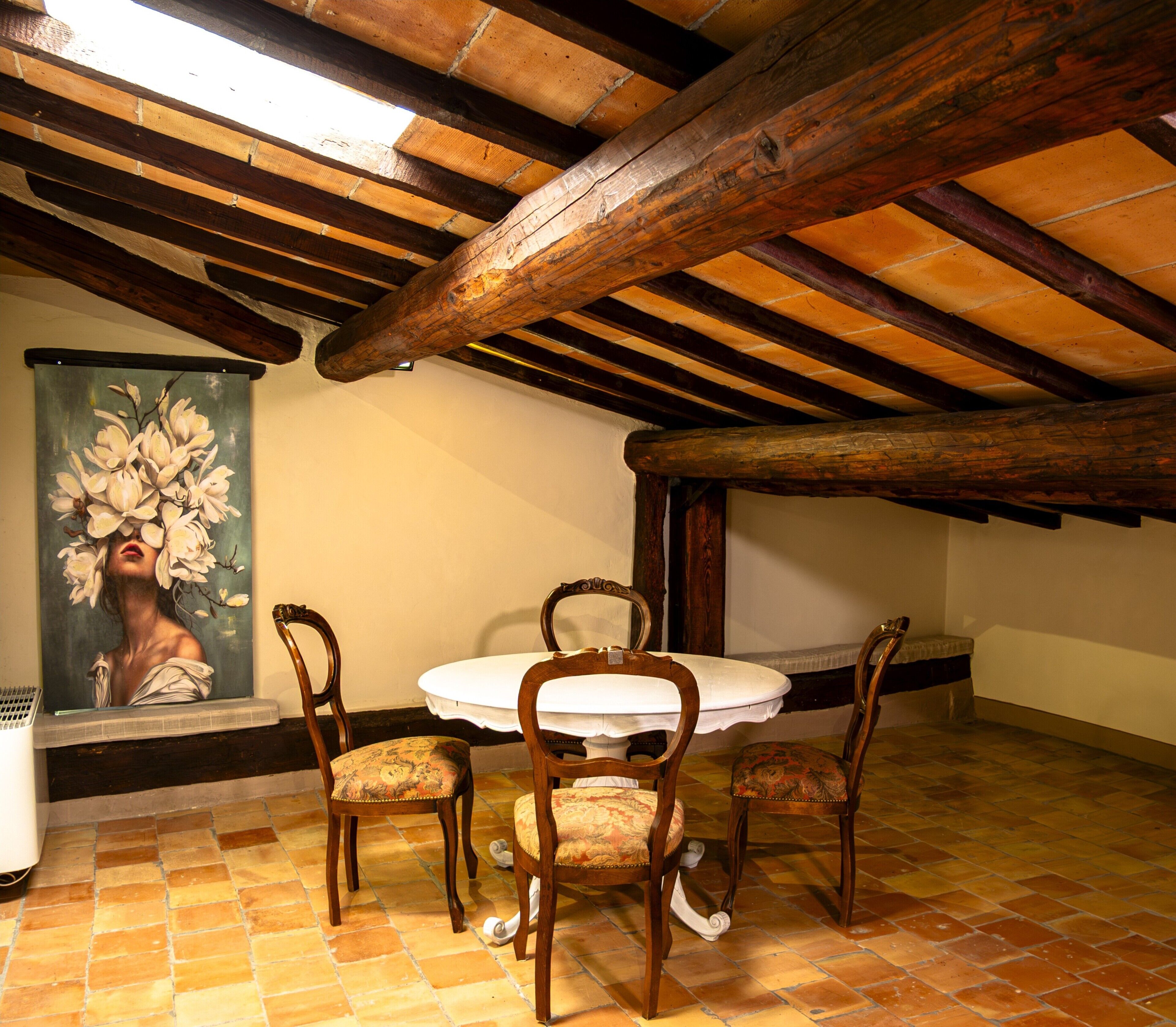 Un comedor con una mesa redonda y cuatro sillas, un cuadro en la pared y vigas de madera a la vista.