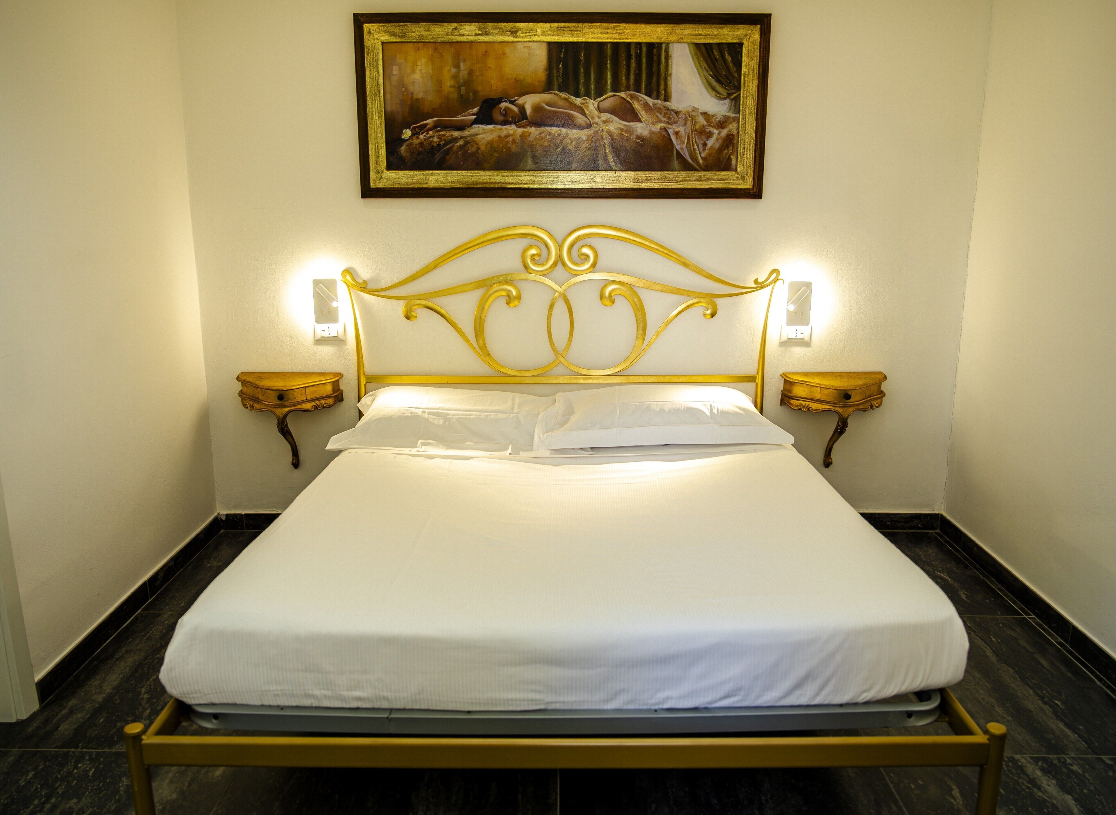 A bed with a gold headboard, white bedding, two bedside tables with lamps, and a painting above the bed.