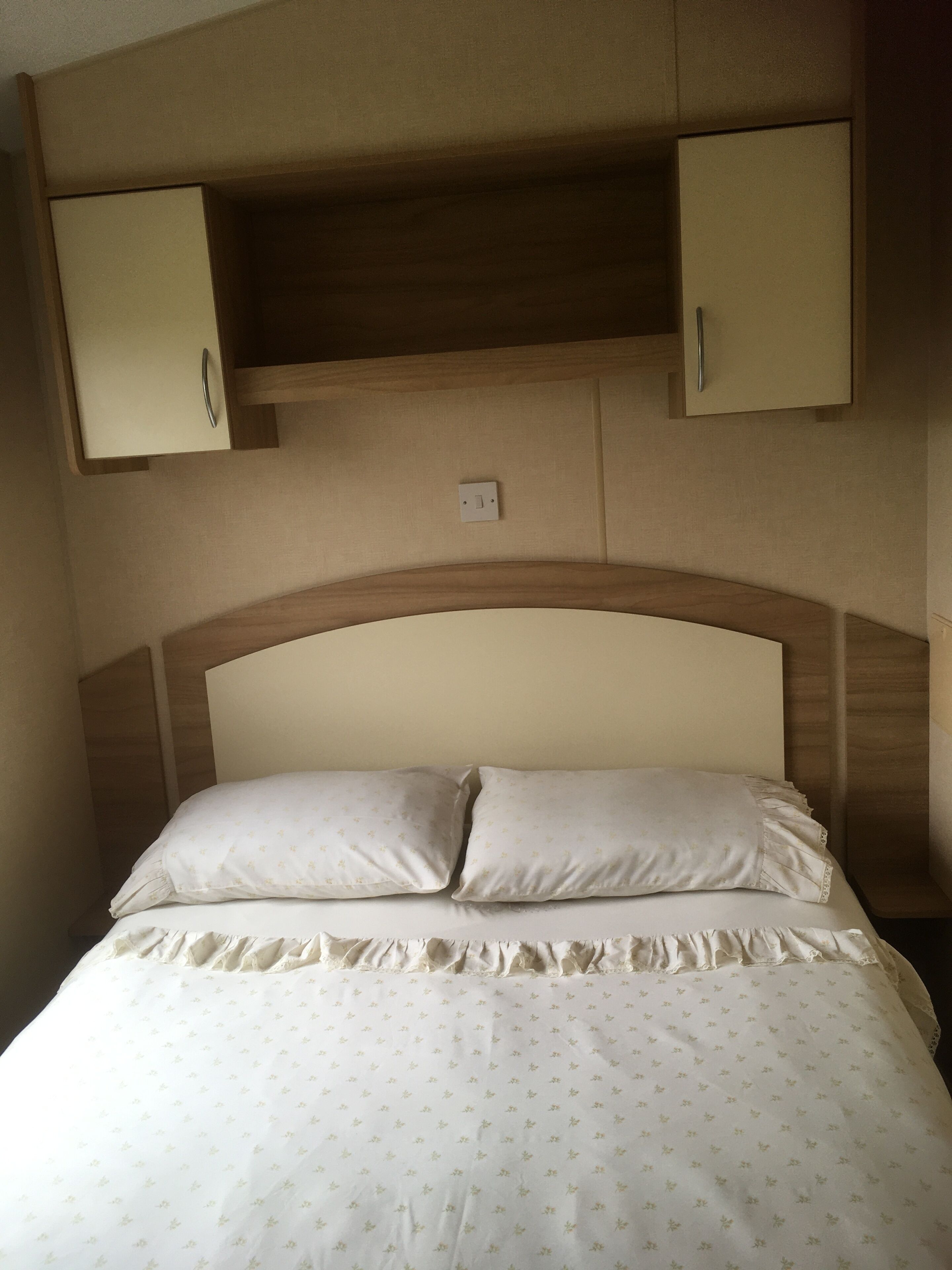 A bed with two pillows, a ruffled bed skirt, and a headboard with a wooden frame. There are two cabinets above the bed, one on each side.