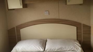 Una cama con dos almohadas, una falda con vuelo y un cabecero con estructura de madera. Hay dos mesitas de noche sobre la cama, una a cada lado.