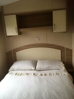 Una cama con dos almohadas, una falda con vuelo y un cabecero con estructura de madera. Hay dos mesitas de noche sobre la cama, una a cada lado.