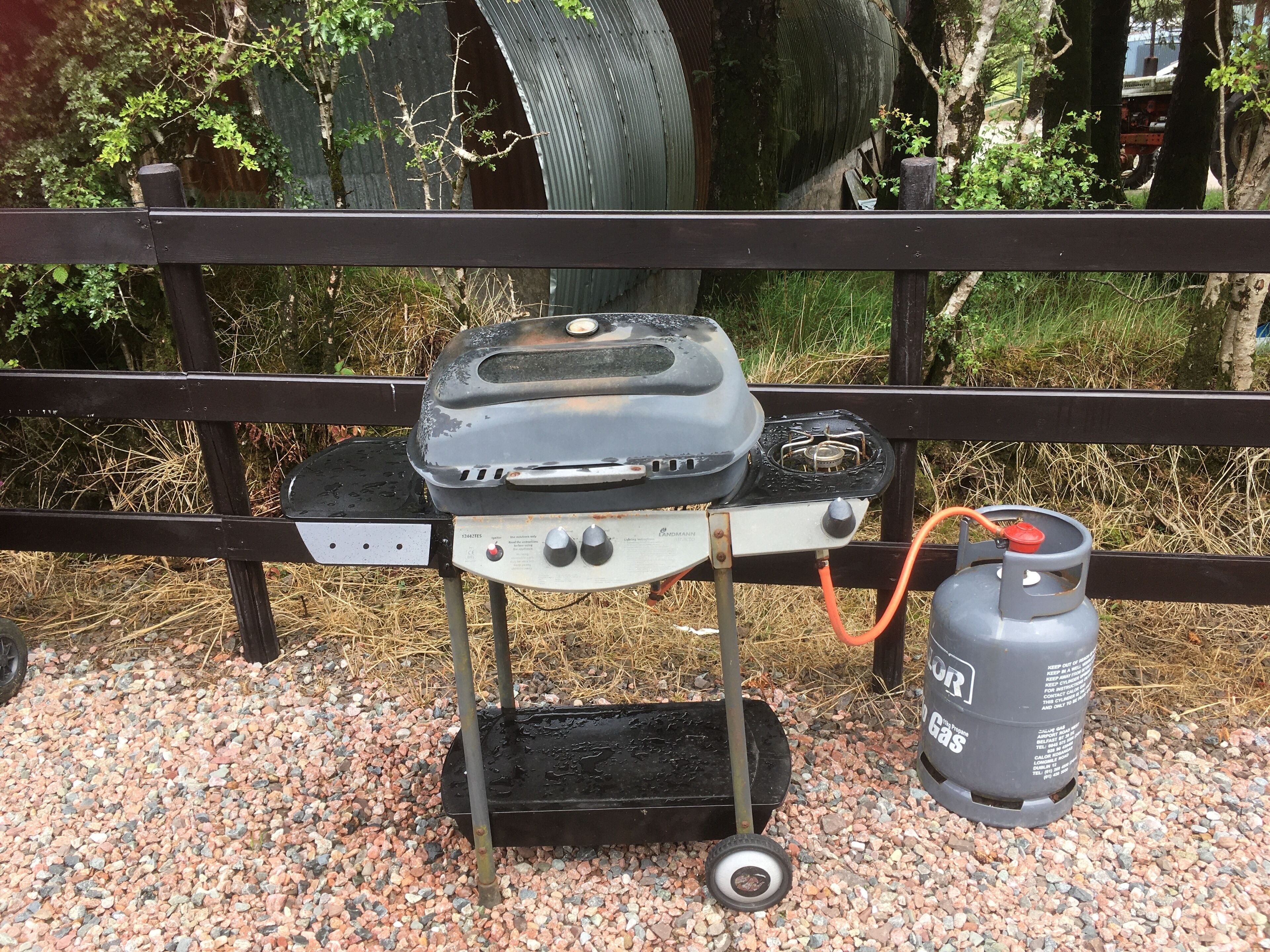 Una parrilla de gas portátil con un cartucho de gas, montada sobre una superficie de grava y un soporte de metal.