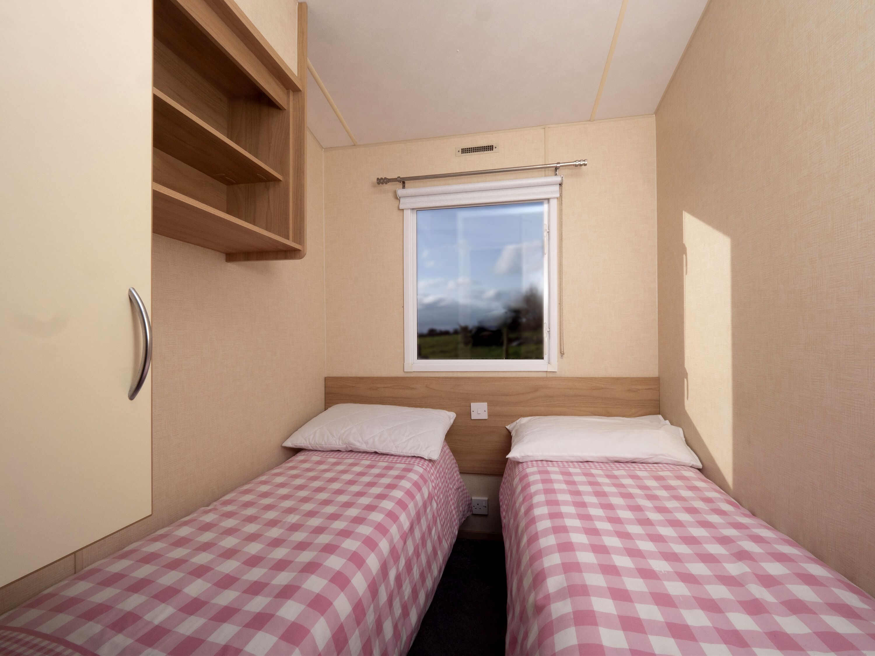 A small room with two single beds, a window with a view of the outdoors, a wooden shelf, and a wall-mounted light switch.
