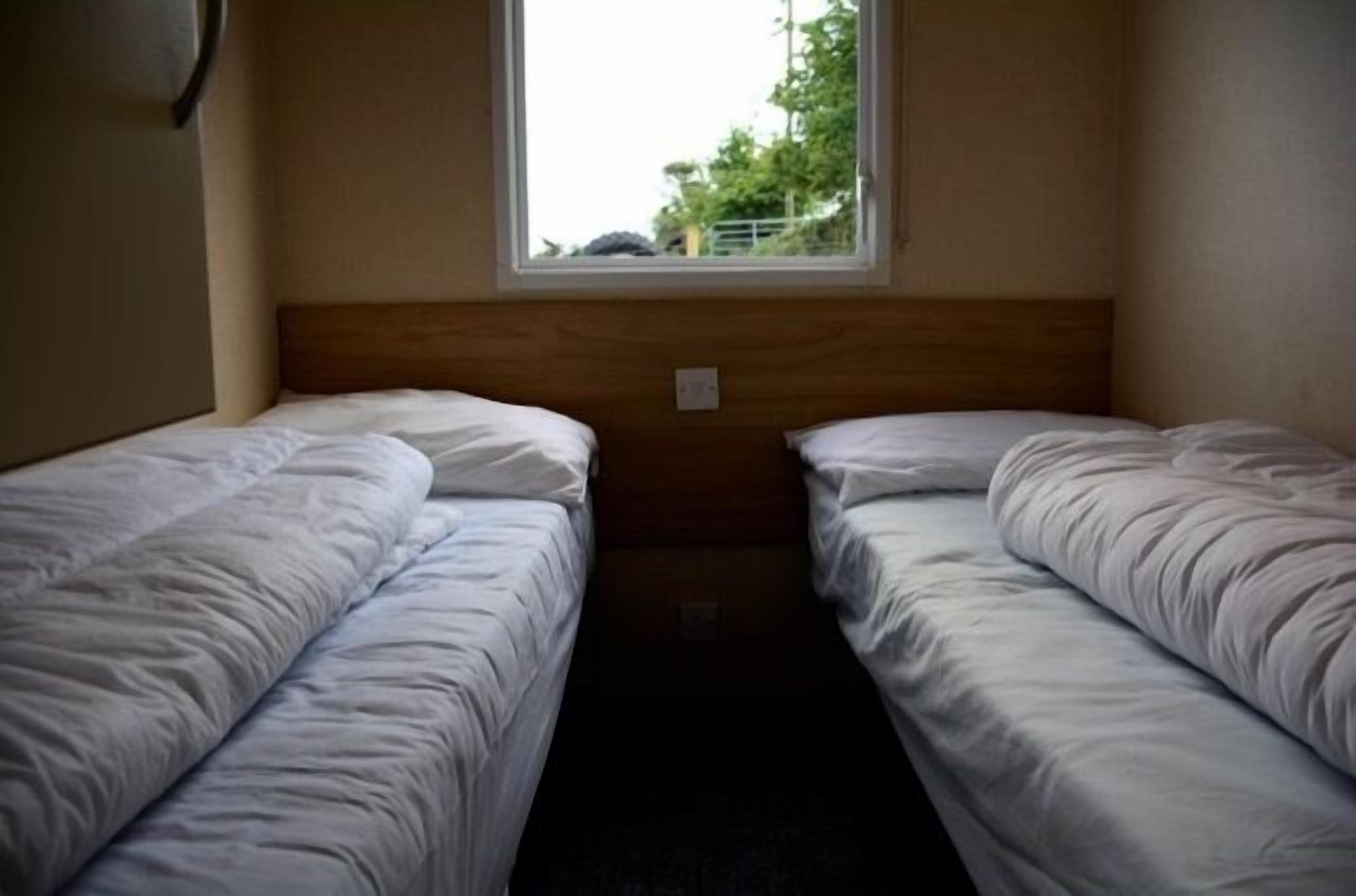 A small, single-room hotel room with two single beds, a wooden headboard, and a window providing a view of greenery outside.