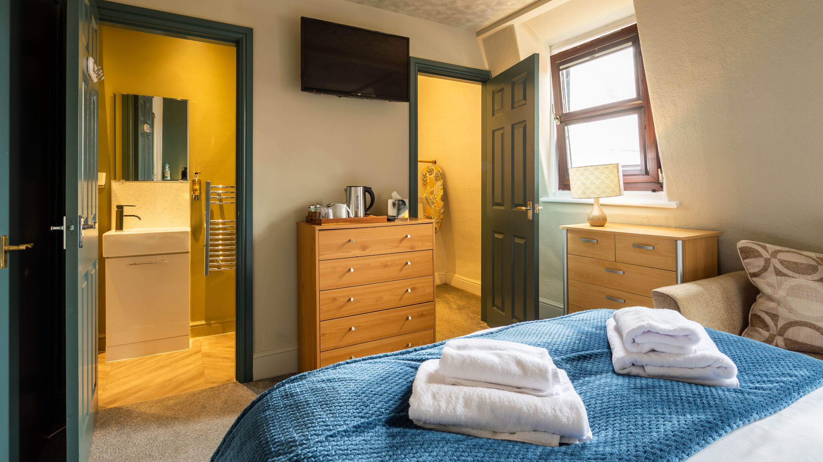 A bedroom with a bed, two dressers, a mirror, a television, and a window.