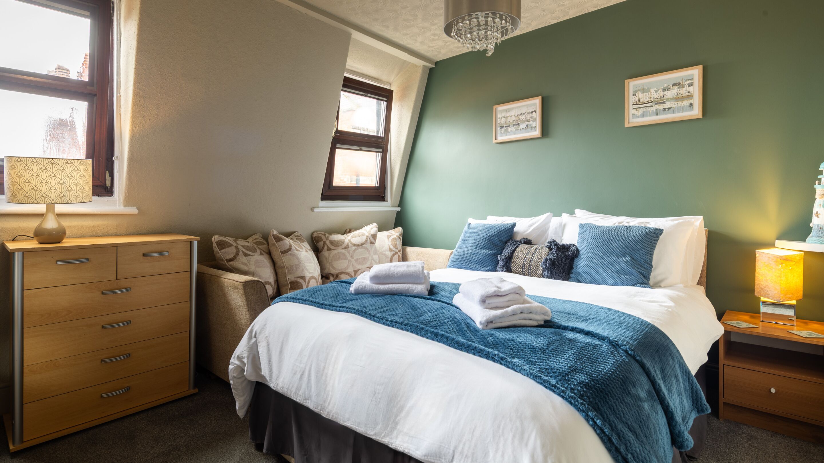 Een slaapkamer met een bed, een dressoir, een nachtkastje, een lamp en twee foto's in lijsten aan de muur.