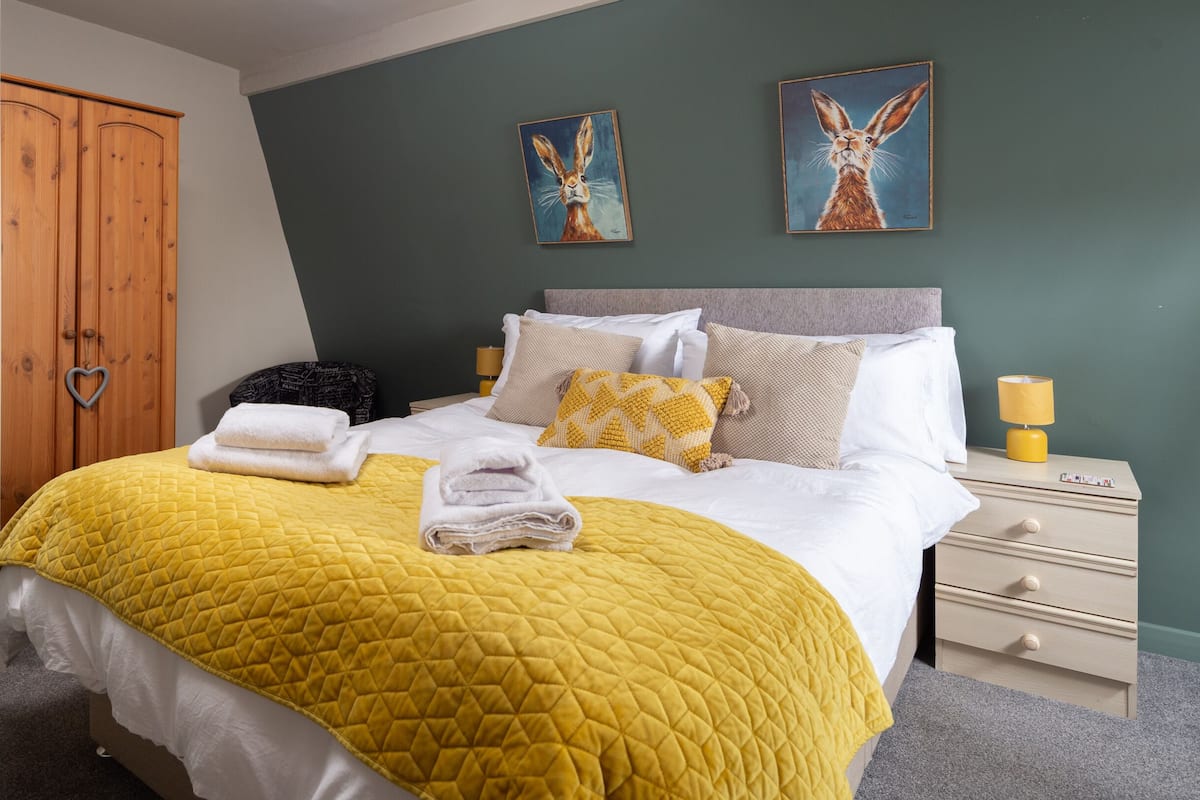 Een keurig opgemaakt bed met een geel sprei, witte kussens en gevouwen handdoeken. Twee schilderijen hangen aan de muur, een houten garderobe en een nachtkastje met een lamp.